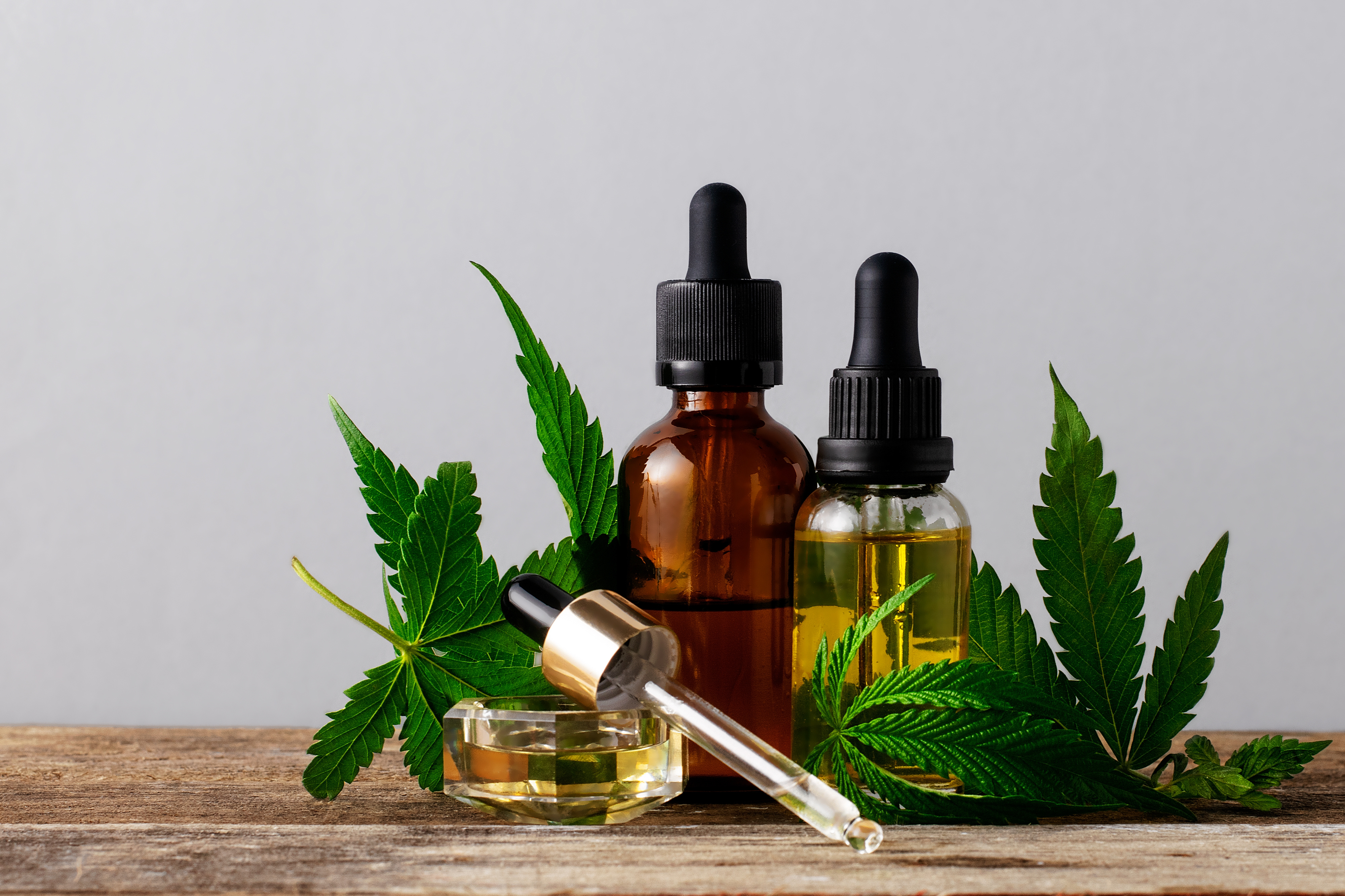 2 small bottles with a droplet laying in front of them, all filled with CBD oil. Small cannabis leaves behind and in front to accent.