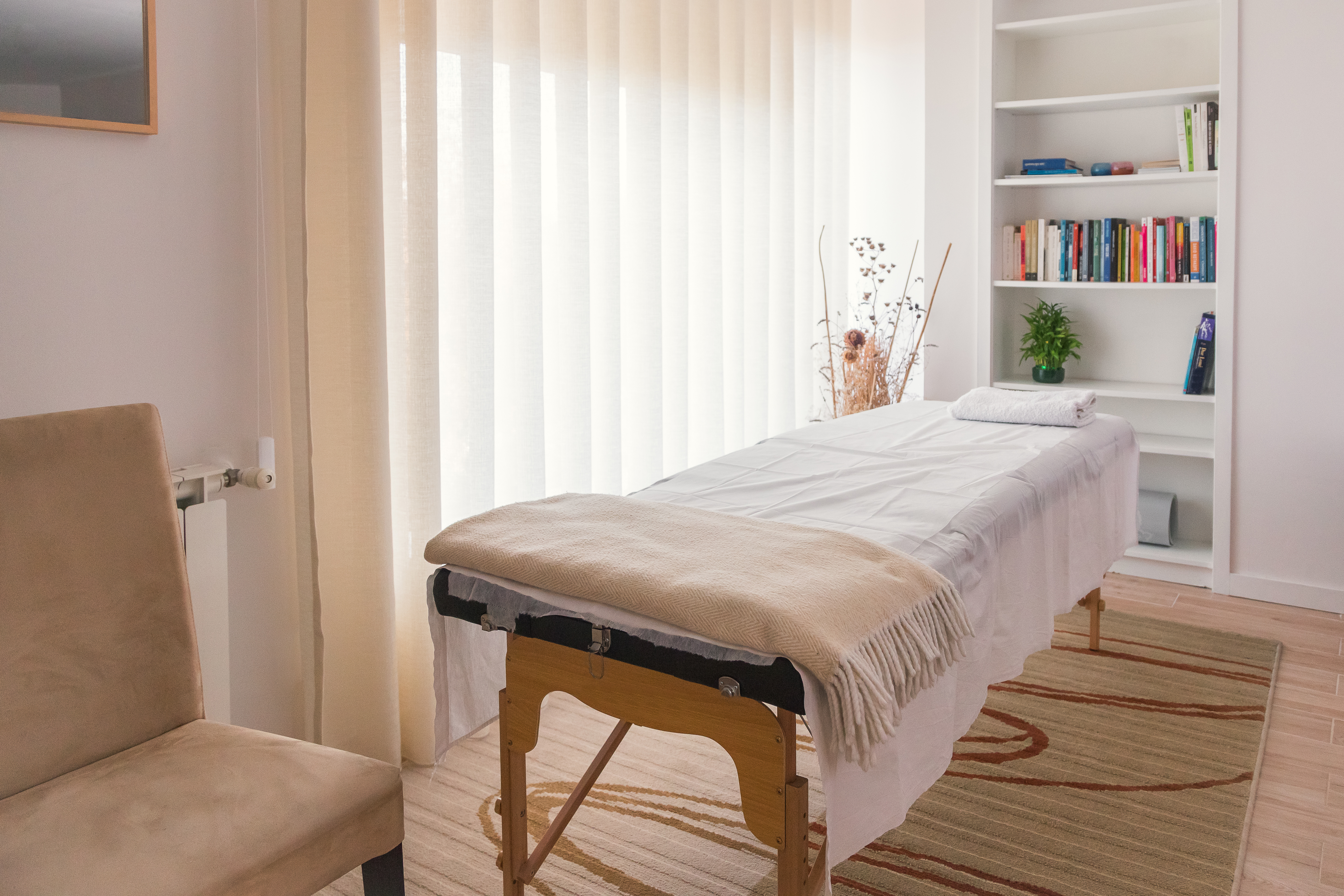 massage room with massage table in middle, bookshelf in the back, sunlight coming through the drapes on the window to the left